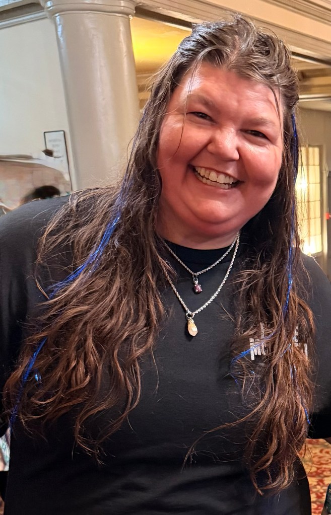 Angela Schneckloth, founder and designer of Warrior Gurl Designs, smiling with her long hair and blue accents visible